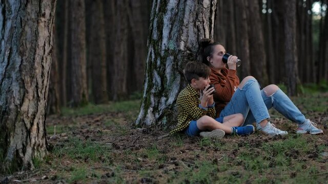 Mother with son relaxing in the forest and drinking hot herbal tea from a thermos on a sunny day. Stay cation 
