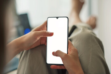 Closeup of a young woman holding a mobile phone with blank white screen sitting at home during the lockdown. Mockup image of a woman using a smartphone in a cozy room.