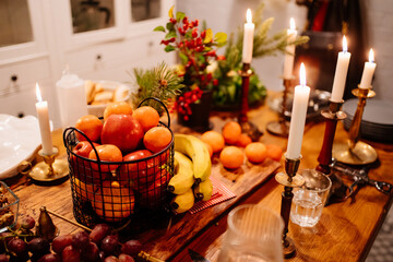Low light. tangerines and apples in a metal basket, burning candles, grapes, bananas, pine branch on the kitchen table. New Year's or Christmas decor. dinner by candlelight in dark. Selective focus.