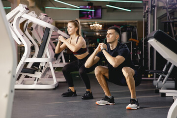 Obraz na płótnie Canvas Two athletes perform squats. The coach shows the correctness of performing the exercises on the feet. Exercise in the gym. Joint training in small groups. A man and a girl train together.