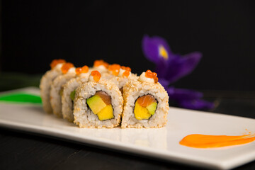 Side view of sushi roll with salmon, avocado, egg omelette wrapped, served with red caviar on top. Iris flower blossom, painting brush stroke decorations on white plate, Black background. Food art
