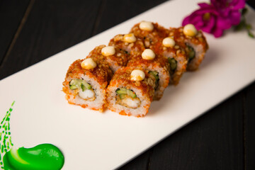 Top view of Japanese sushi roll with tempura black tiger shrimp, cucumber served with sauce. Purple flower blossom and green painting brush stroke decoration on white plate, Black Wooden background
