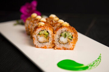 Close up to crunchy Japanese sushi roll with tempura black tiger shrimp, cucumber served with sauce. Purple flower blossom and green painting brush stroke decoration on white plate, Black background
