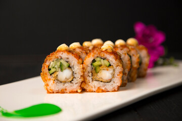 Close up to crunchy Japanese sushi roll with tempura black tiger shrimp, cucumber served with sauce on top. Purple flower blossom and green painting brush stroke decoration on white plate. Food art
