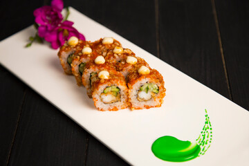 Side view of Japanese sushi roll with tempura black tiger shrimp, cucumber served with sauce. Purple flower blossom and green painting brush stroke decoration on white plate, Black Wooden background
