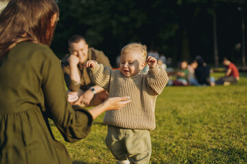 cute little caucasian boy learning to walk on lawn in park. His mother opening wide arms to catch him. Father on background, watching with tenderness.  Image with selective focus