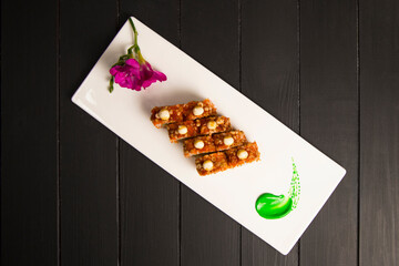 Top view of crunchy Japanese sushi roll served with sauce. Purple flower blossoms and green painting brush stroke decoration on rectangular white plate. Directly above shot. Black Wooden background
