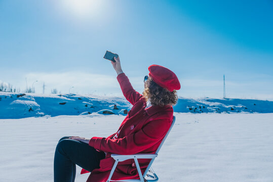 A woman dressed in red with a hat sitting on a chair makes a selfie in a totally snowy fiel