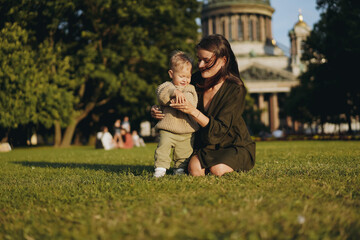 Young Caucasian lady, mother of little son sitting on grass in front of st isaac’s cathedral and helping boy stand holding him by hand. Image with selective focus