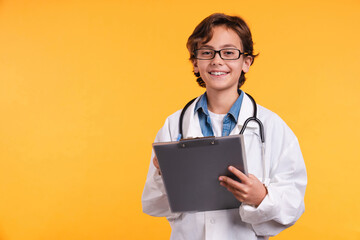 Cheerful little boy future doctor in medical clothes with stethoscope and clipboard isolated over yellow background