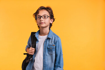 Smart little white boy with backpack in glasses like a pupil isolated over yellow background