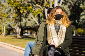 Fototapeta na wymiar Woman sitting on a park bench with her back to a man with surgical mask