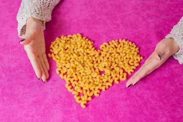Pink background. A heart is made of pasta. Women's hands.