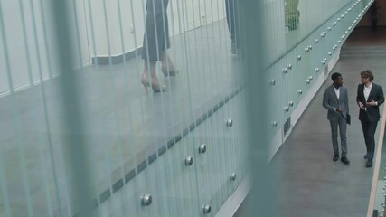 Wide shot of young African-American and Caucasian businessmen wearing elegant suits going along business center hall and having chat