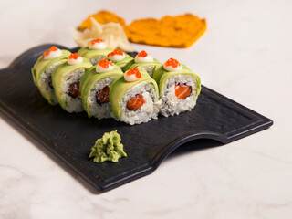 Close up to Japanese sushi roll with avocado and red caviar on top served with wasabi and ginger on black plate, heart shapes of turmeric powder spices on white marble background. Salmon inside roll
