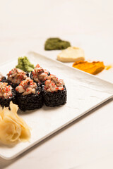 Side view of Japanese black rice sushi roll with unagi eel served with ginger, wasabi on square plate. Heart shapes if basil, garlic, turmeric powder spices on white marble background
