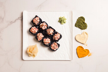 Obraz na płótnie Canvas From above of Japanese black rice sushi roll with unagi eel served with ginger, wasabi on square plate. Heart shapes if basil, garlic, turmeric powder spices on white marble background 