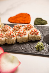 Japanese sushi roll with roasted salmon and tobiko flying fish roe on top served on black plate with wasabi and ginger. heart shapes of turmeric and basil spice, rose petals out of focus on foreground