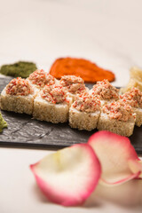 Japanese sushi roll with roasted salmon and tobiko flying fish roe on top served on black plate with wasabi and ginger. heart shapes of turmeric and basil spice, rose petals out of focus on foreground