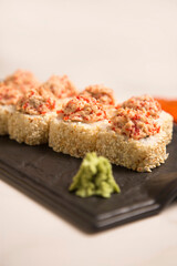 Close up to sushi roll with roasted salmon and tobiko flying fish roe on top wrapped in rice, sesame seeds served on black plate with wasabi on marble background

