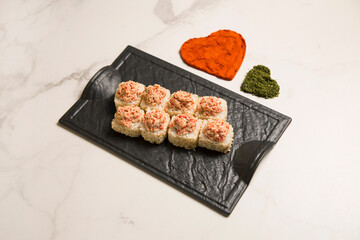 Close up of Japanese sushi roll with roasted salmon and tobiko flying fish roe on top served on black plate. heart shapes of turmeric and basil spices on marble background. Asian menu dish
