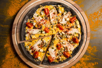 Italian black dough pizza served on wooden board. Slice of meat pizza with chicken, tomatoes, blue cheese, champions and melted Gouda cheese. Black chalkboard with scattered turmeric spices
