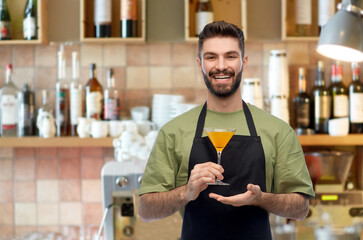 alcohol drinks, people and profession concept - happy smiling barman in black apron holding orange martini cocktail glass over bar background