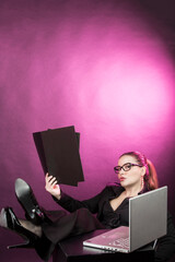 Manager donna con i capelli raccolti  con occhiali da vista, vestita con  tailleur nero lavora al computer con i piedi sopra il tavolo , isolata su sfondo rosso