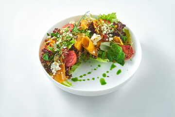 Appetizing and dietetic salad of roasted pumpkin, arugula, feta and tomatoes in the white plate on a white background.
