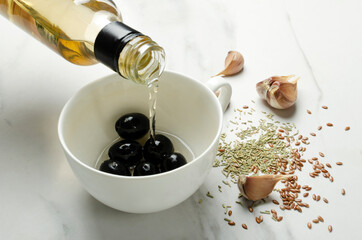 Closeup of pouring herbal oil nto the whie cup, black olives, herbs on the white table