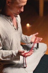 Young attractive man in natural clothes for practice sits in lotus position in a dark room for meditation with rosary in his hands
