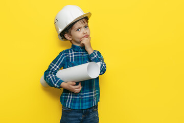 Serious little kid boy engineer or architect in a protective helmet holding construction plan over yellow background.