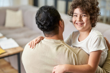 Fototapeta premium Feeling protected by dad. Playful latin boy having fun while spending time with his father, hugging him while sitting in the living room at home