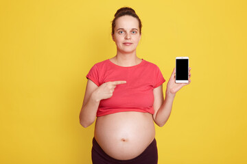Young pregnant woman with pleasant appearance showing phone with blank screen while standing...