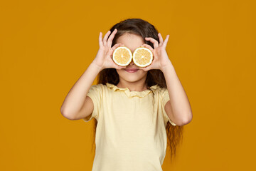 Obraz na płótnie Canvas cute little child girl holding fresh juicy lemon against her eyes isolated over yellow background