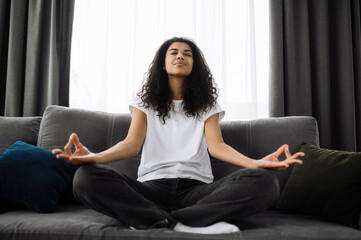 Calm young woman sits on sofa at home and chilling. Beautiful african american woman is practise yoga and meditation in lotus position. Cute smiling girl relieve stress, calmness concept