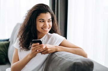 Online communication. Happy african american girl, holds her smartphone, thoughtfully looks away and smiling. Young woman use her gadget while sitting on a couch browsing internet, chatting with