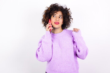 young beautiful caucasian woman wearing pink knitted sweater against white wall speaks on mobile phone spends free time indoors calls to friend.