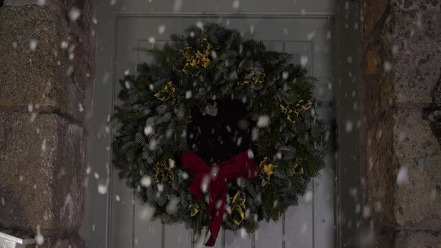 4K: Christmas wreath hung on a front door and it's snowing. The snow falls outside the house at night. Stock Video Clip Footage