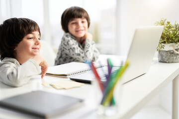 Two smiling little hispanic boys, twin brothers looking at the laptop screen during online lesson for children, sitting at the table at home