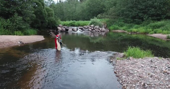 girl in red cloak crosses river into ford, carries sticks, branches, firewood in her hands. Walking over stones along river bank with a waterfall. Forest, evening. Fabulous image, fairytale character.