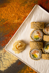 Directly above of Japanese Tempura hot sushi roll served with wasabi, ginger on white square plate. Dark background with scattered spices: dried garlic, turmeric, curry, paprika powder
