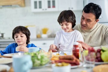 Loving middle aged hispanic father serving his cute little son, sitting with kids at kitchen table while having a breakfast at home