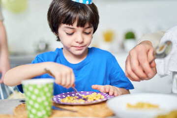 Birthday boy. Adorable little latin american boy in birtday cap eating while celebrating birthday together with his family at home