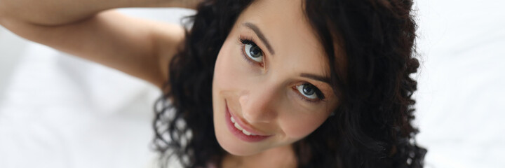 Obraz premium Top view of cute brunette female posing on camera in bed. Lovely smiling lady with pretty wavy haircut. Person in good mood. Rest at home. Morning and wake up concept