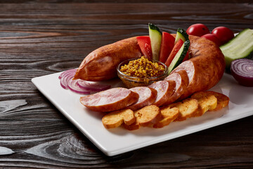 Sausages with a garnish of vegetables and herbs with sauces and bread on a white rectangular plate on a dark wooden table.