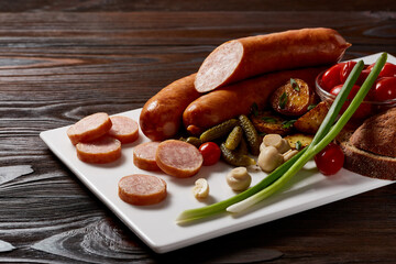 Sausages with a garnish of vegetables and herbs with sauces and bread on a white rectangular plate on a dark wooden table.