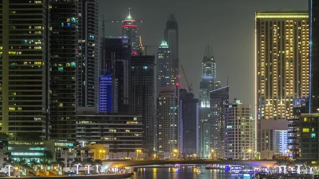Promenade and bridge with traffic over canal in Dubai Marina timelapse at night, UAE. View from bridge with palms, floating boats and illuminated towers. District with artificial canal