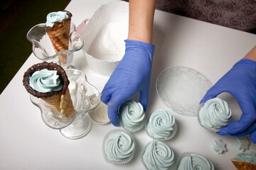 Woman in rubber gloves makes marshmallows. Nearby is a zephyr in a waffle cone. Dessert lies on the table surface.