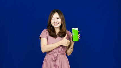beautiful asian woman pointing green screen in handphone isolated blue background